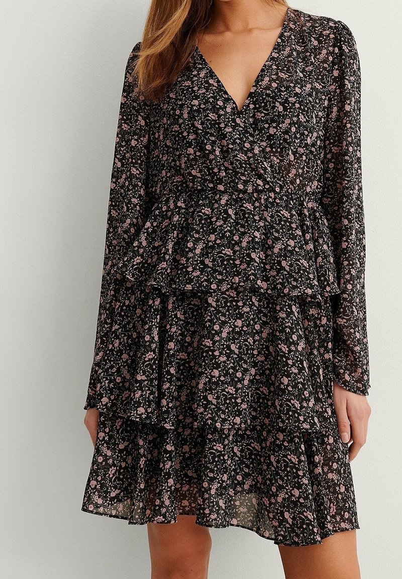 Femme portant une robe portefeuille noire à fleurs avec manches longues et jupe à volants superposés, debout devant un mur clair uni.