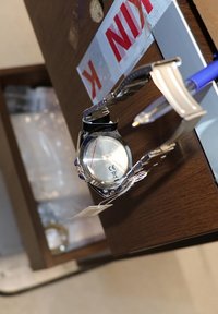 Silberne Edelstahl-Armbanduhr mit rundem Zifferblatt, wasserdichtem Gehäuse und schwarzem Armband. Enthält ein angehängtes Etikett für Informationen.