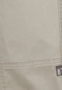 Beige stoff med en jevn tekstur, med synlige sømmer og en liten etikett på høyre side. Materialet ser holdbart og lett ut.