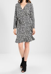 Vestido negro ajustado con un patrón floral blanco, mangas largas, un cinturón que se ata en la cintura y un dobladillo con volantitos. Llevado con botas negras hasta el tobillo.