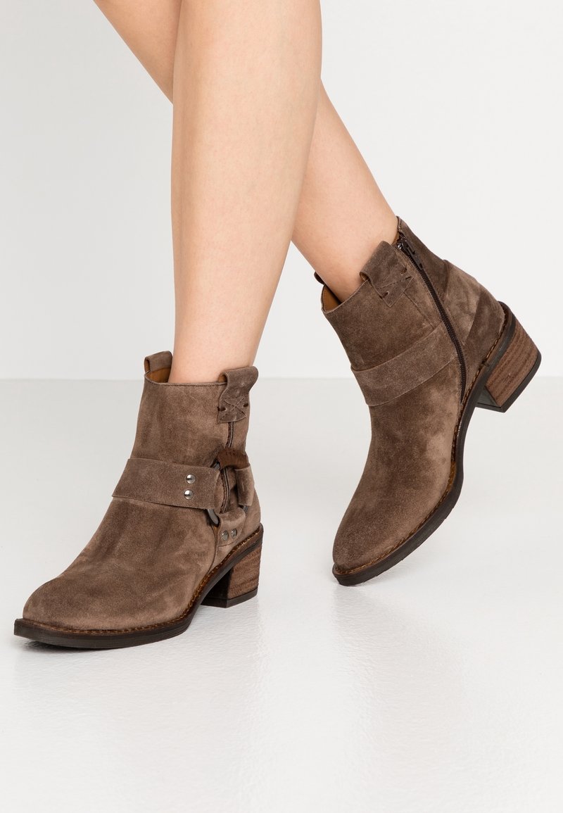 Botas de tobillo de ante marrón con un tacón grueso, cremallera lateral, correa decorativa con acentos metálicos y un diseño de puntera ligeramente puntiaguda.