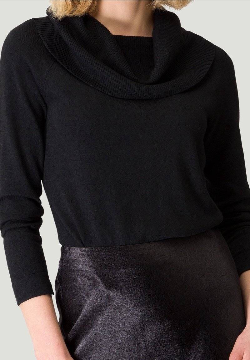 Femme portant un pull noir à col châle à manches longues associé à une jupe en satin noir brillant, coupée au niveau du milieu du torse.