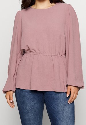 Woman wearing a mauve long-sleeve blouse with elastic waist and blue jeans, standing against a plain light background.