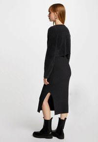 Zwart, cropped sweater van pluche materiaal, gecombineerd met een aansluitende, geribbelde zwarte midi-jurk met een split aan de zijkant. Draagt zwarte enkellaarzen.