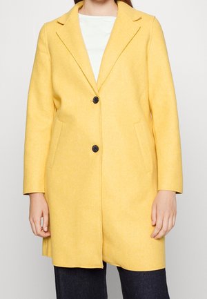 Kvinde iført en gul frakke i mellem længde med revers og to sorte knapper, kombineret med mørke bukser og en hvid top.