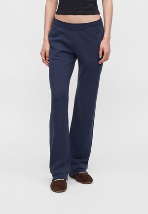 Persona con pantaloni della tuta blu scuro a gamba larga e scarpe marroni slip-on, in piedi su uno sfondo chiaro e uniforme.