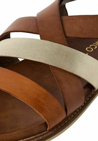 Sandale en cuir brun et beige avec des brides croisées. Elle présente des bandes texturées et une semelle plate avec un intérieur marron clair.