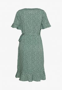 Robe verte clair jusqu'aux genoux avec des pois noirs, manches courtes volantées, ourlet froncé et ceinture nouée à la taille, vue de dos.