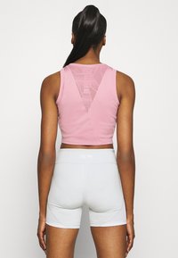 Mujer con cabello largo y oscuro en una coleta, que lleva una camiseta de tirantes rosa corta con detalle de malla y pantalones cortos deportivos de cintura alta en gris claro.