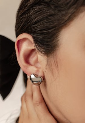 Herzförmige Silberstecker mit glänzender Metalloberfläche, die eine glatte, reflektierende Oberfläche zeigen. Am Ohr getragen, mit schlichtem Design.