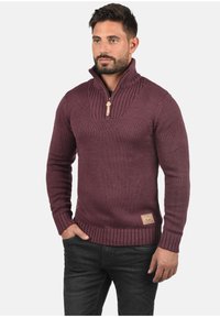 Maglione lavorato a maglia color bordeaux con colletto alto e zip, polsini e orlo a costine, e un motivo testurizzato. Etichetta sul lato sinistro in basso.