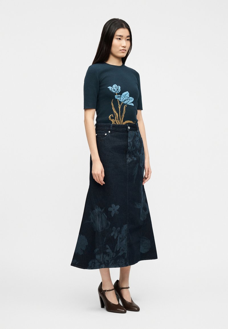 Vrouw draagt een donkerblauw kort mouwshirt met blauw bloemmotief en een donkerblauwe maxi rok met subtiele bloempatronen, gecombineerd met bruine hakken.