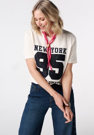 Lachende vrouw met blond haar draagt een wit New York 95 t-shirt, rode nek sjaal en blauwe jeans met studs aan de zijkant, poseert tegen een witte achtergrond.