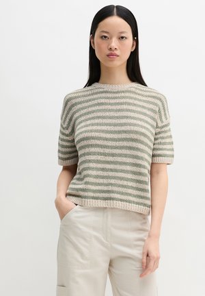 Donna con capelli neri lisci che indossa un maglione a righe verde e beige a maniche corte e pantaloni beige, in piedi con una mano in tasca.