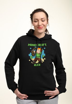 Žena v černé mikině s kapucí s nápisem "Minecraft Alex" a pixelovanou postavou držeící luk, stojící před jednoduchým světlým pozadím.