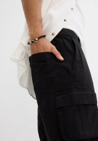 Pantaloni cargo neri con tasche frontali, abbinati a una camicia bianca con texture. Un braccialetto marrone con chiusura in argento adornano il polso.