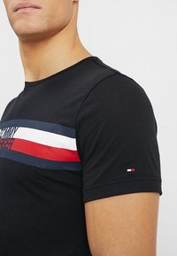 Černé bavlněné tričko s horizontálním červeným, bílým a modrým pruhovaným designem na hrudi, s logem a malým logem na rukávu.