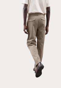 Pantalones beige de corte recto, con bolsillos laterales y traseros, y una textura suave, acompañados de zapatos marrón oscuro.
