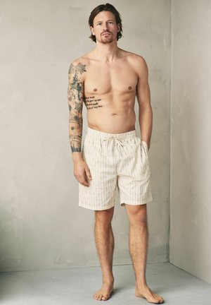 Shirtless man with tattoos on left arm and chest wearing cream striped shorts, standing barefoot against a plain light wall.