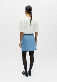 Femme aux cheveux tressés portant un t-shirt blanc surdimensionné, une mini-jupe en denim bleu, des collants noirs et des mocassins noirs, de dos sur un fond uni.
