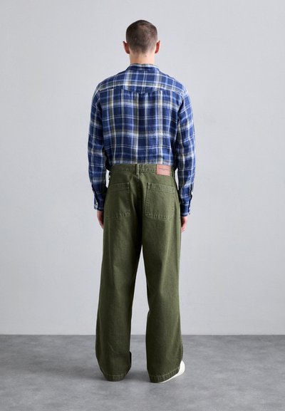 Καρό πουκάμισο flannel σε μπλε αποχρώσεις, συνδυασμένο με ανοιχτόχρωμες παντελόνες σε ελαιόχρωμο που έχουν πίσω τσέπες και καφέ δερμάτινο λογότυπο.