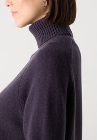 Tumši purpursarkans turtlenecks džemperis, kas izgatavots no mīksta adīšanas materiāla, ar ribotām detaļām ap kaklu un brīvā fitā.