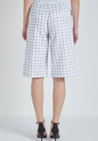 Pantalones cortos blancos a cuadros con patrón de rejilla azul marino, corte entallado, hasta la rodilla, con dos bolsillos traseros, combinados con zapatos de tacón negro.