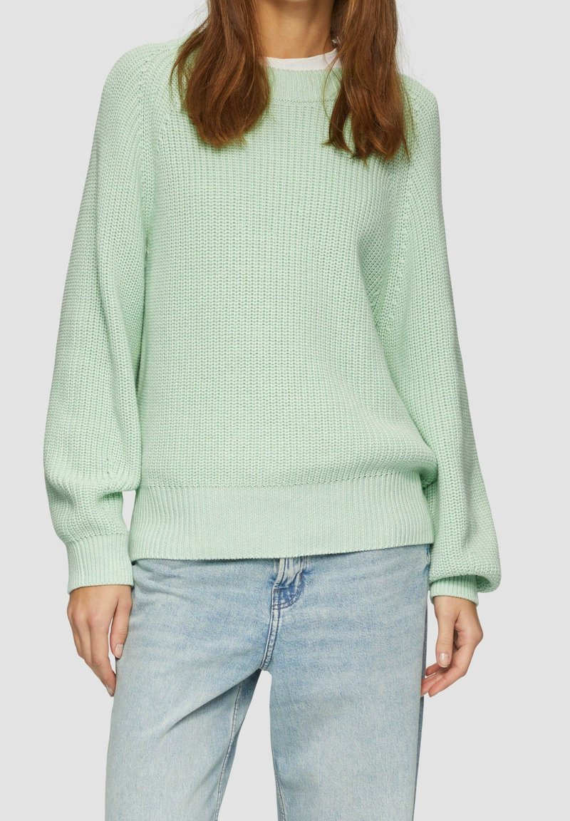 Femme portant un pull vert clair en maille et un jean bleu clair, debout devant un fond uni et clair.