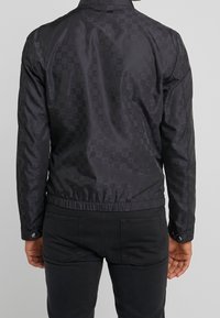 Svart rutigt klickå med en slank, glänsande material och elastisk midja. Har knäppte ärmar och subtila logotypdetaljer.