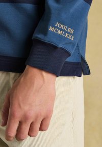 Nahaufnahme einer Hand neben beigen Cordhosen, die ein blaues Langarmshirt trägt, auf dem "JOULES MCMLXXX" nahe dem Bündchen gestickt ist.