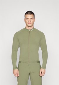 Haut de sport à manches longues, couleur vert olive, avec une fermeture éclair sur le devant, présentant un design ajusté et une légère texture, porté avec un bas de sport assorti.