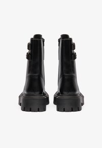 Botas de cuero negras hasta el tobillo con suela de goma gruesa, que cuentan con dos hebillas laterales y un acabado suave. El diseño es robusto y estilizado.