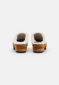 Zoccoli in legno marrone con foderatura in faux fur bianco, tacco basso, cuciture a vista e suola in gomma. Design open-back per una facile calzata.