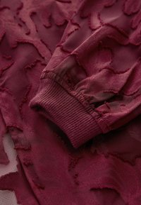 Tissu bordeaux texturé avec un motif floral en relief, doté d'un poignet côtelé. Le matériau semble doux et transparent avec des détails complexes.