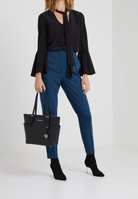 Czarna bluzka z długim rękawem i rozszerzanymi mankietami, niebieskie dopasowane spodnie oraz czarna torba na ramię ze złotymi detalami. Model nosi czarne botki za kostkę.