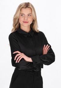 Sort bluse med lange ærmer i glat stof med spidst krave, knappet front og manchetærmer, stylet med sorte bukser i taljen.