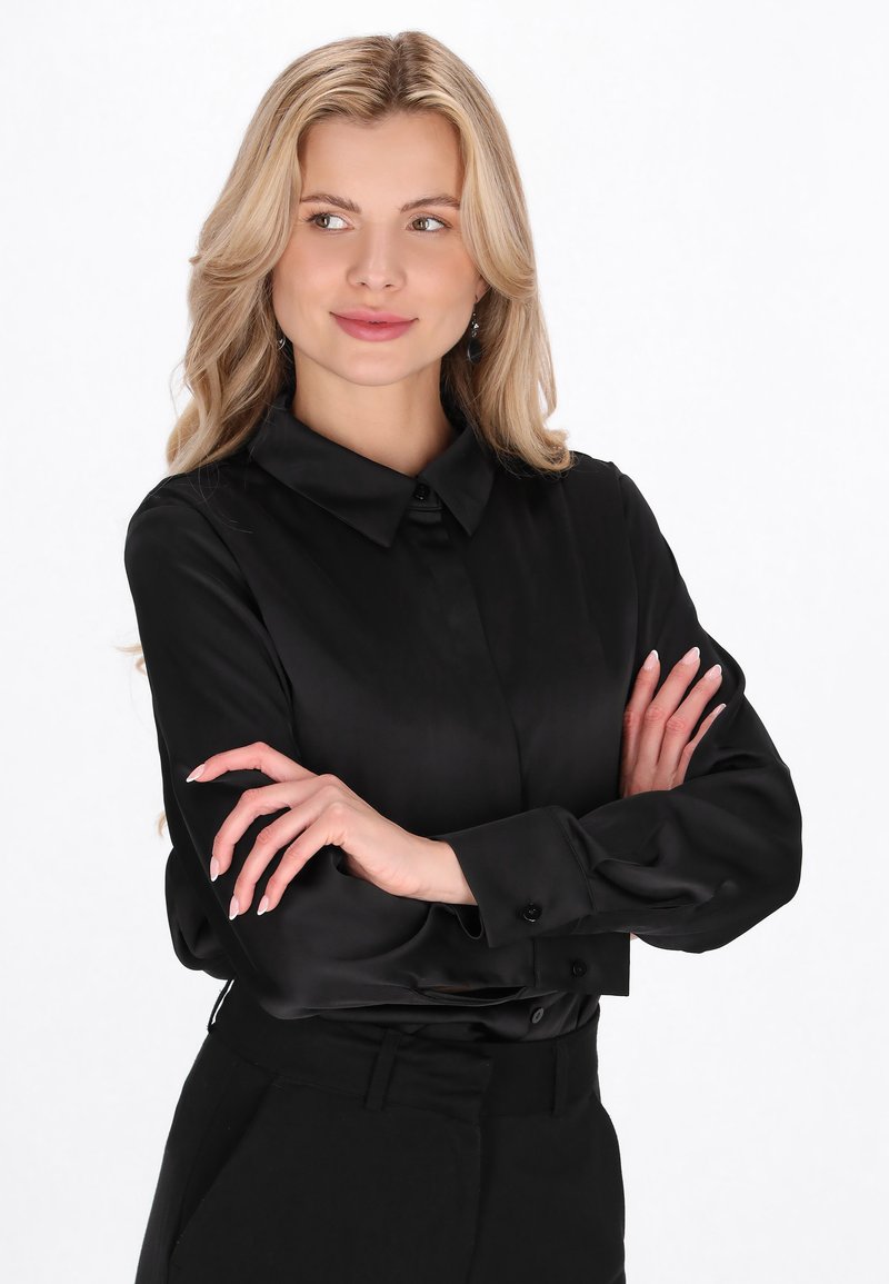 Sort bluse med lange ærmer i glat stof med spidst krave, knappet front og manchetærmer, stylet med sorte bukser i taljen.