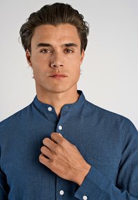 Camicia blu con bottoni, colletto alzato, realizzata in tessuto testurizzato. Presenta una tasca sul petto e bottoni bianchi, mostrando un primo piano di una mano che regola il colletto.