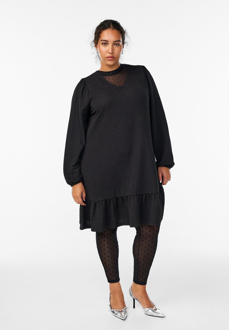 Kvinde med krøllet hår iført en sort langærmet kjole over polkaprikkede mesh leggings og sølvfarvede spidse højhælede sko, stående mod en hvid baggrund.