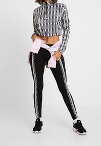 Černá krátká bunda s vertikálním bílým logem, doplněná černými legínami s bílými postranními pruhy a černými teniskami.