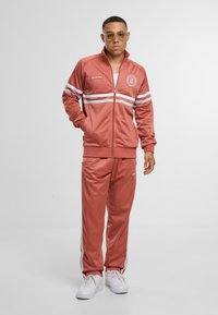 Træningsdragt i blødt, rust-rødt stof med hvide striber, lynlåsjakke og elastiske manchetter. Sammen med hvide sneakers. Accentueret logo på brystet.