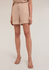 Pantaloni corti in lino beige con vita con cintura, orli risvoltati e una texture liscia, abbinati a sandali con tacco nude e con cinturini.