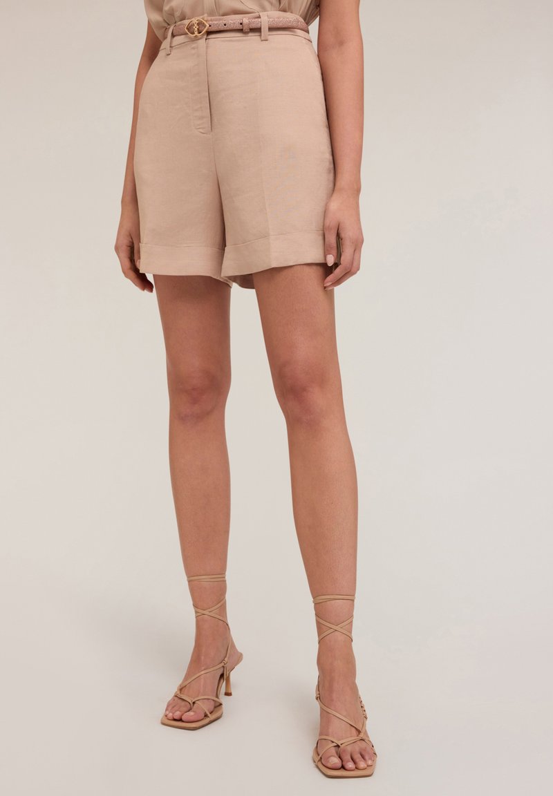 Pantaloni corti in lino beige con vita con cintura, orli risvoltati e una texture liscia, abbinati a sandali con tacco nude e con cinturini.