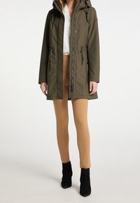 Olijfgroene parka met capuchon, ritssluiting en knoopsluiting, trekkoorddetails en een gladde textuur. Draagbaar over beige leggings en zwarte enkellaarsjes.