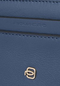 Primo piano di pelle blu con testo in rilievo "PIQUADRO" e piccolo logo in metallo dorato sotto la cucitura.