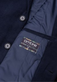 Manteau bleu foncé avec boutons noirs, ouvert pour montrer la doublure bleu marine lisse et l'étiquette de marque "ombre QUINTESSENCE" taille M.