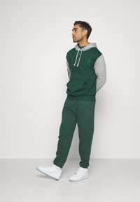Pantalones de chándal verdes y sudadera a juego con mangas grises, con "NEW YORK" impreso en verde oscuro. Zapatillas blancas, estilo casual, textura suave.