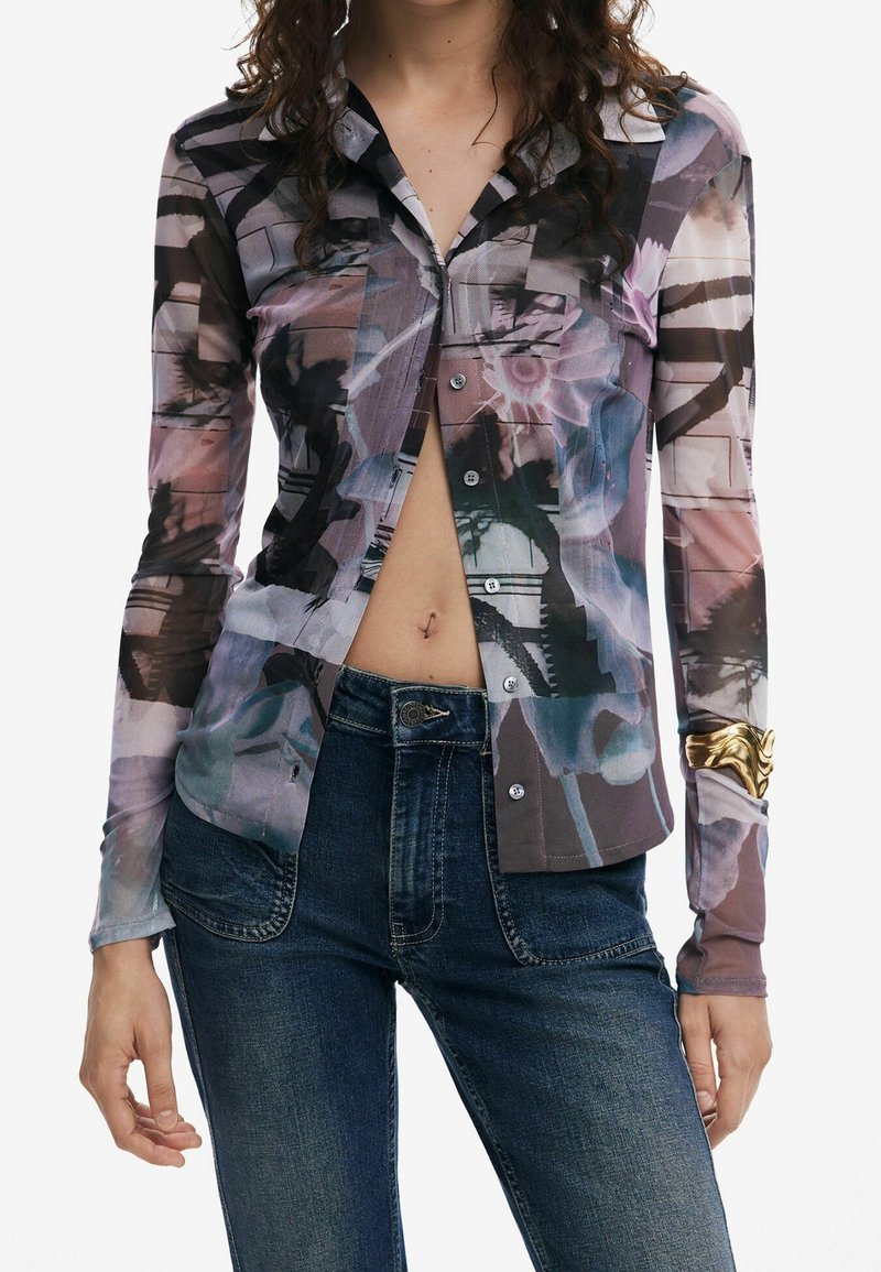 Femme portant une chemise à manches longues partiellement déboutonnée à motif abstrait, un jean bleu et un bracelet en or au poignet gauche.