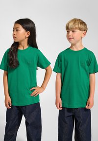 Två barn som bär gröna bomulls t-shirts med ribbad halsringning, i kombination med mörkblå denimbyxor. T-shirtsen har en liten logotyp på bröstet.