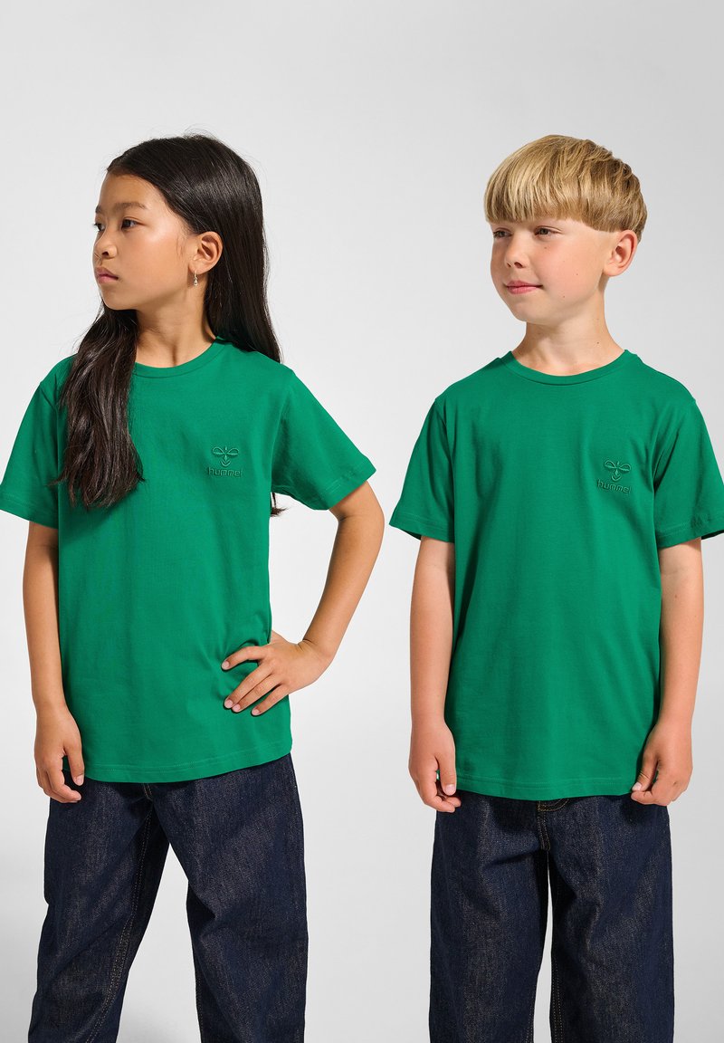 Två barn som bär gröna bomulls t-shirts med ribbad halsringning, i kombination med mörkblå denimbyxor. T-shirtsen har en liten logotyp på bröstet.